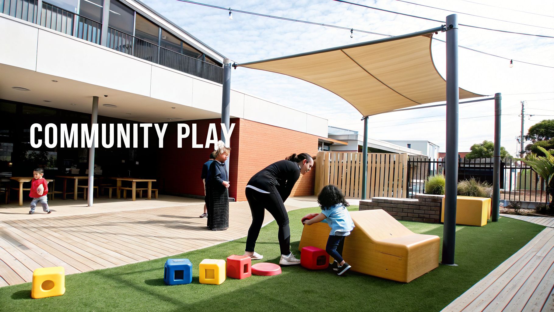 An outdoor community play area with children, adults, colorful soft play equipment on artificial grass, and a wooden deck.